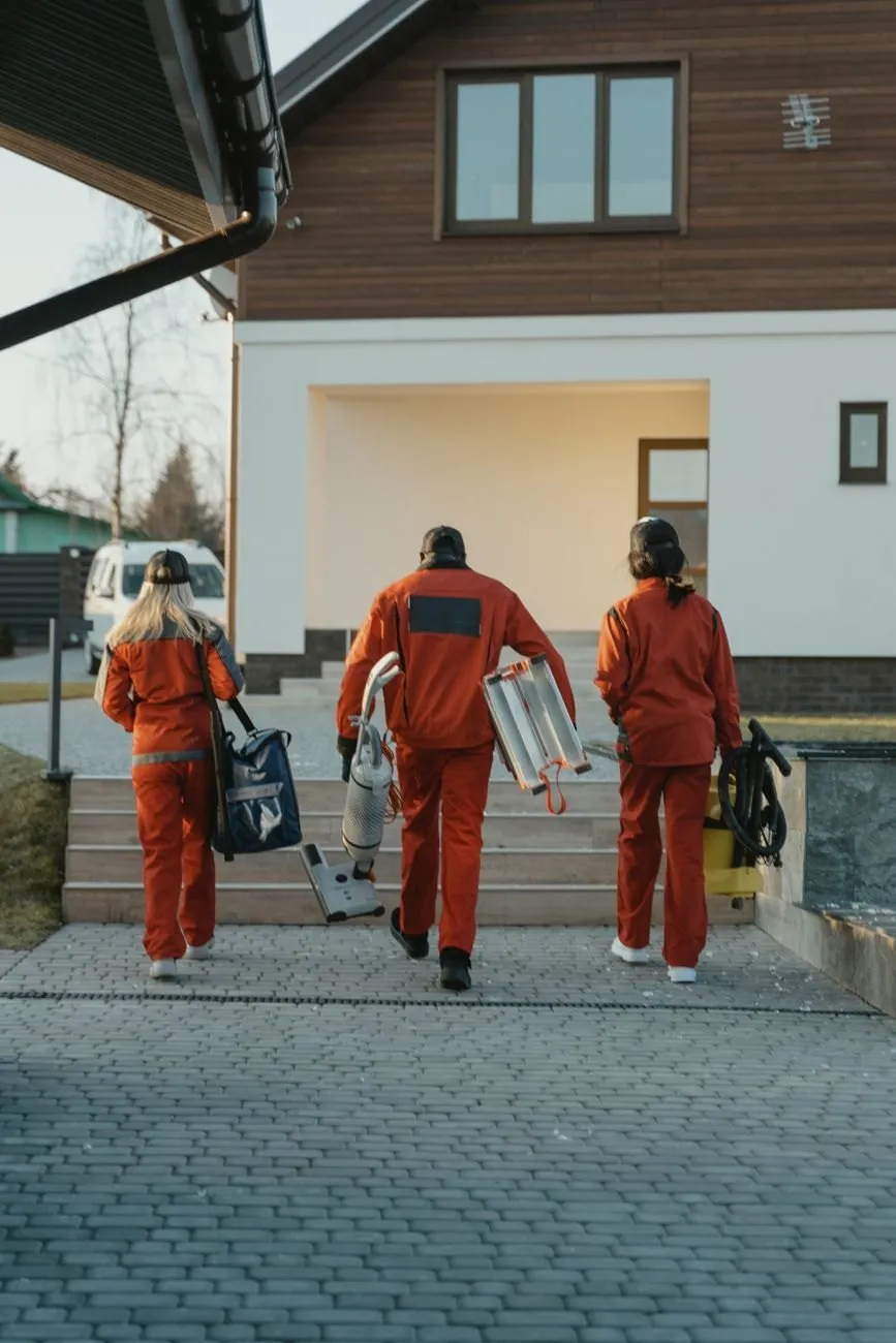 Three Briskwell cleaners seen from behind, carrying a vacuum, a step ladder and kit bags up to a house