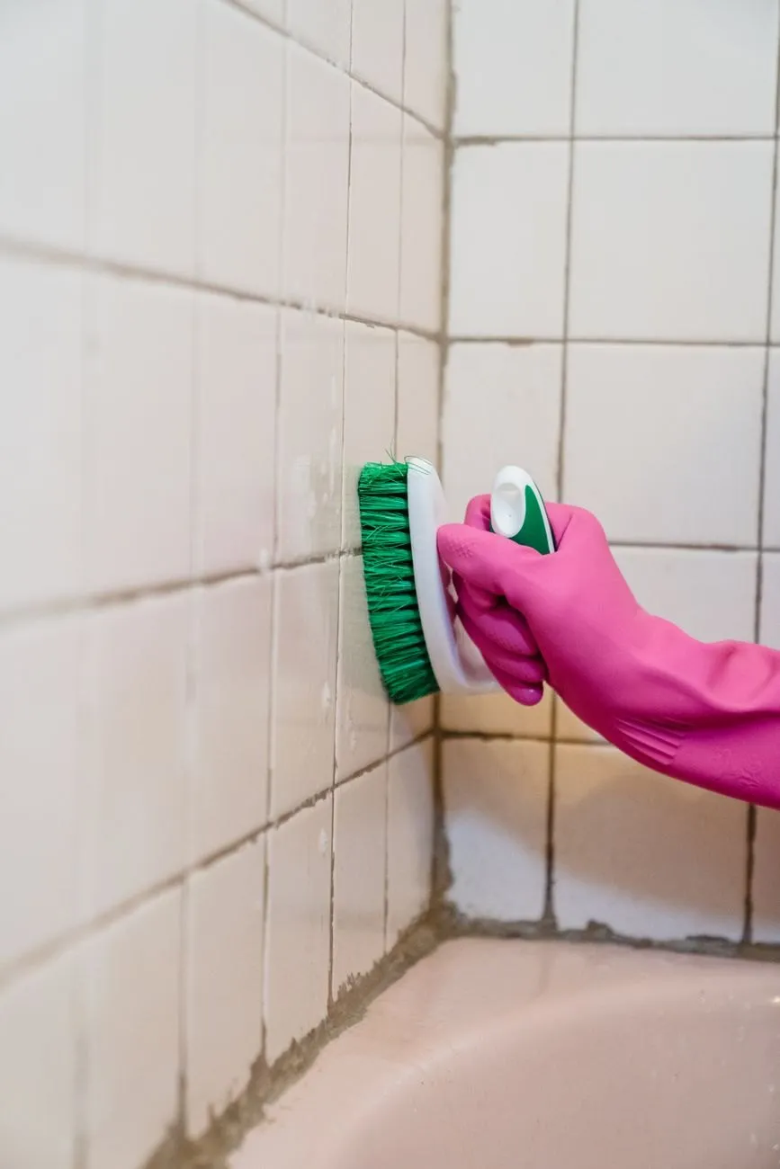 A pink-gloved hand scrubbing tile grout with a small brush