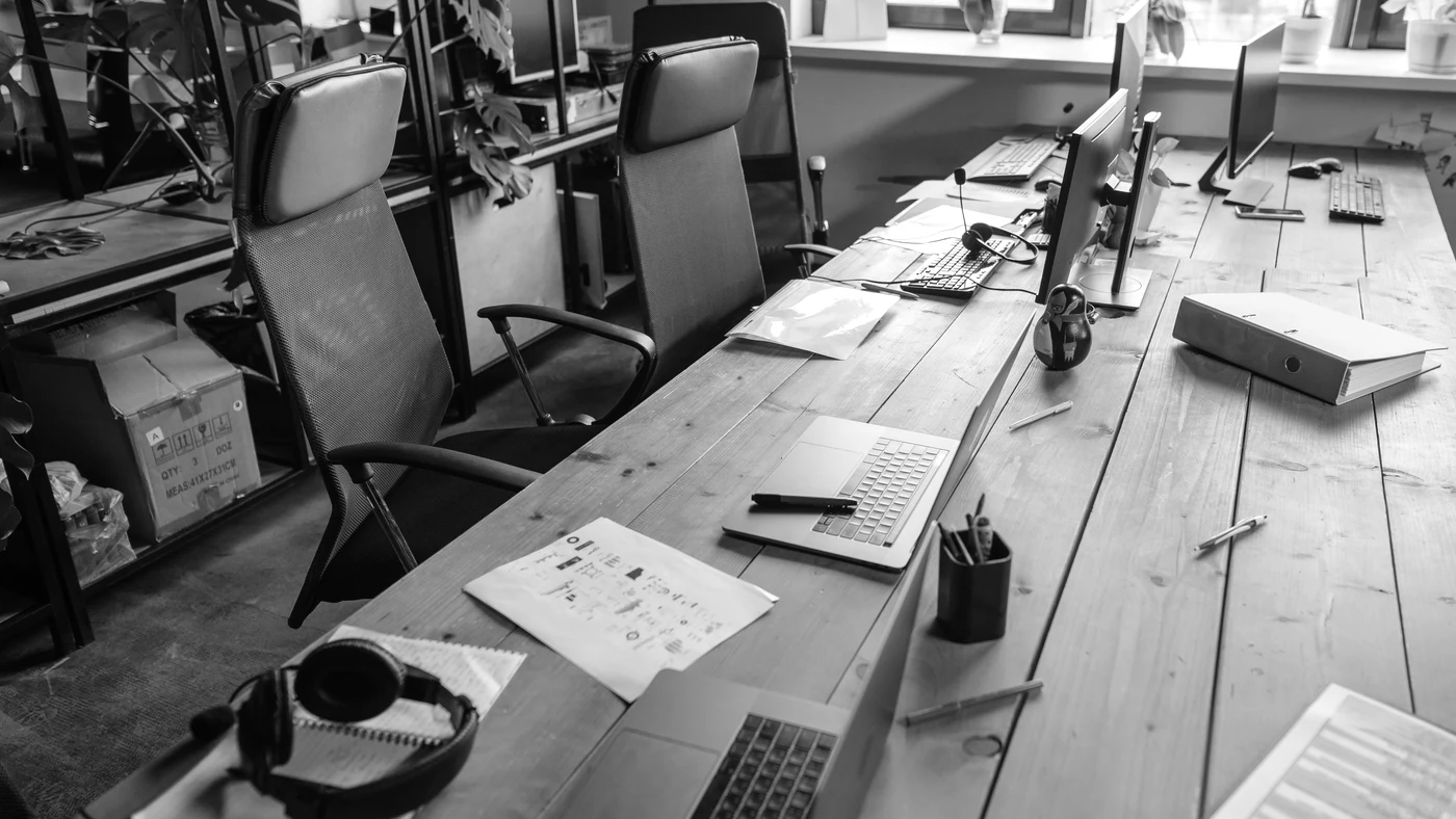An open-plan office of long shared desks, printed achromatic with the specular return at full.